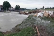 Genangan banjir mengalir di area permukiman warga dengan tanggul darurat dan material kayu berserakan di tepi aliran air akibat luapan sungai.