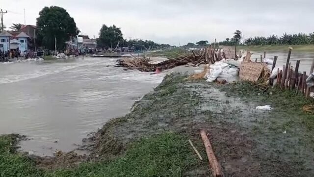 Genangan banjir mengalir di area permukiman warga dengan tanggul darurat dan material kayu berserakan di tepi aliran air akibat luapan sungai.