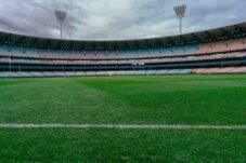Stadion sepak bola kosong dengan tribun penonton dan lapangan rumput hijau di bawah langit mendung.