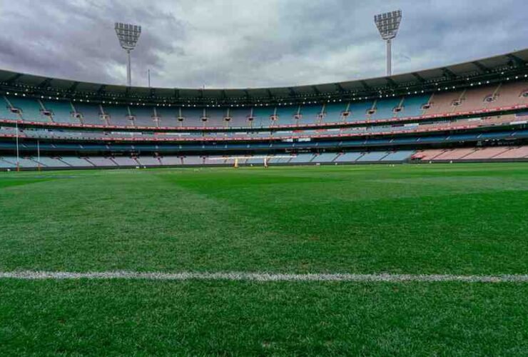 Stadion sepak bola kosong dengan tribun penonton dan lapangan rumput hijau di bawah langit mendung.